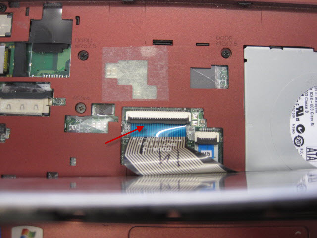 Arrow points to brown catch that needs gently lifting to release the keyboard ribbon cable.